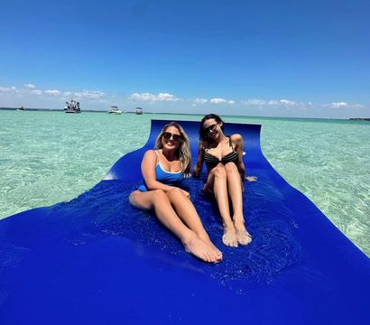Two smiling women in swimsuits lounging on a bright blue floating mat in shallow turquoise water with anchored boats on the horizon and a clear sunny sky