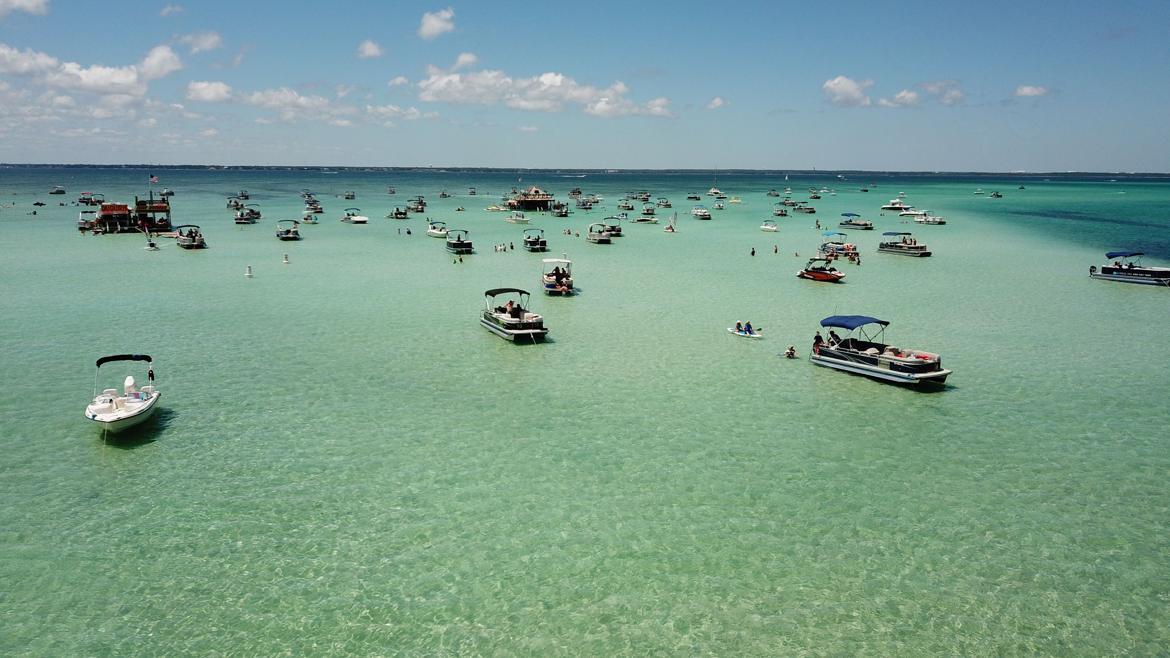 Aerial view of a turquoise shallow bay dotted with anchored pontoon and motorboats, paddleboarders and swimmers enjoying a sunny blue-sky day