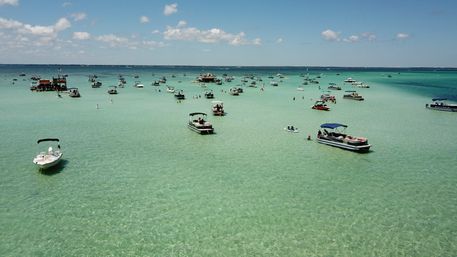 Aerial view of a turquoise shallow bay dotted with anchored pontoon and motorboats, paddleboarders and swimmers enjoying a sunny blue-sky day