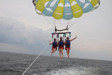 Three people parasailing over the open ocean, suspended from a yellow-and-teal parachute and towline, wearing life jackets against a cloudy sky