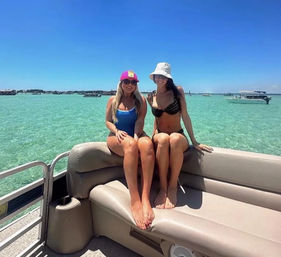 Two women in swimsuits and sun hats sitting on a pontoon boat bench with clear turquoise water, scattered boats on the horizon, and a bright blue sky on a sunny day