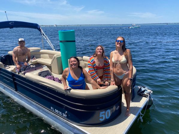 Group of four friends in swimsuits smiling on a navy pontoon boat with a tall teal float, enjoying a sunny day on calm coastal waters.