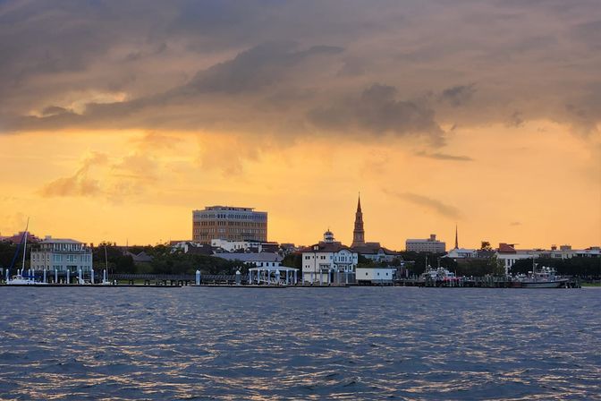 Golden sunset over a coastal city waterfront skyline with church steeples and low‑rise buildings, docks and moored boats under dramatic clouds, rippling harbor water in the foreground.