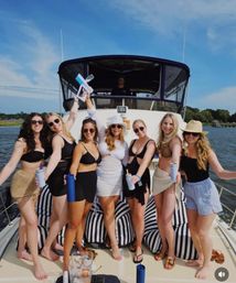 Cheerful group of friends in swimsuits on the bow of a yacht with striped cushions, holding drinks and a water gun under a bright blue sky over calm coastal waters.