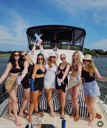 Cheerful group of friends in swimsuits on the bow of a yacht with striped cushions, holding drinks and a water gun under a bright blue sky over calm coastal waters.