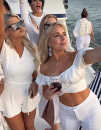 Group of women in white outfits on a yacht at sunset, cheering and taking a selfie while one holds a glass of red wine with the ocean in the background
