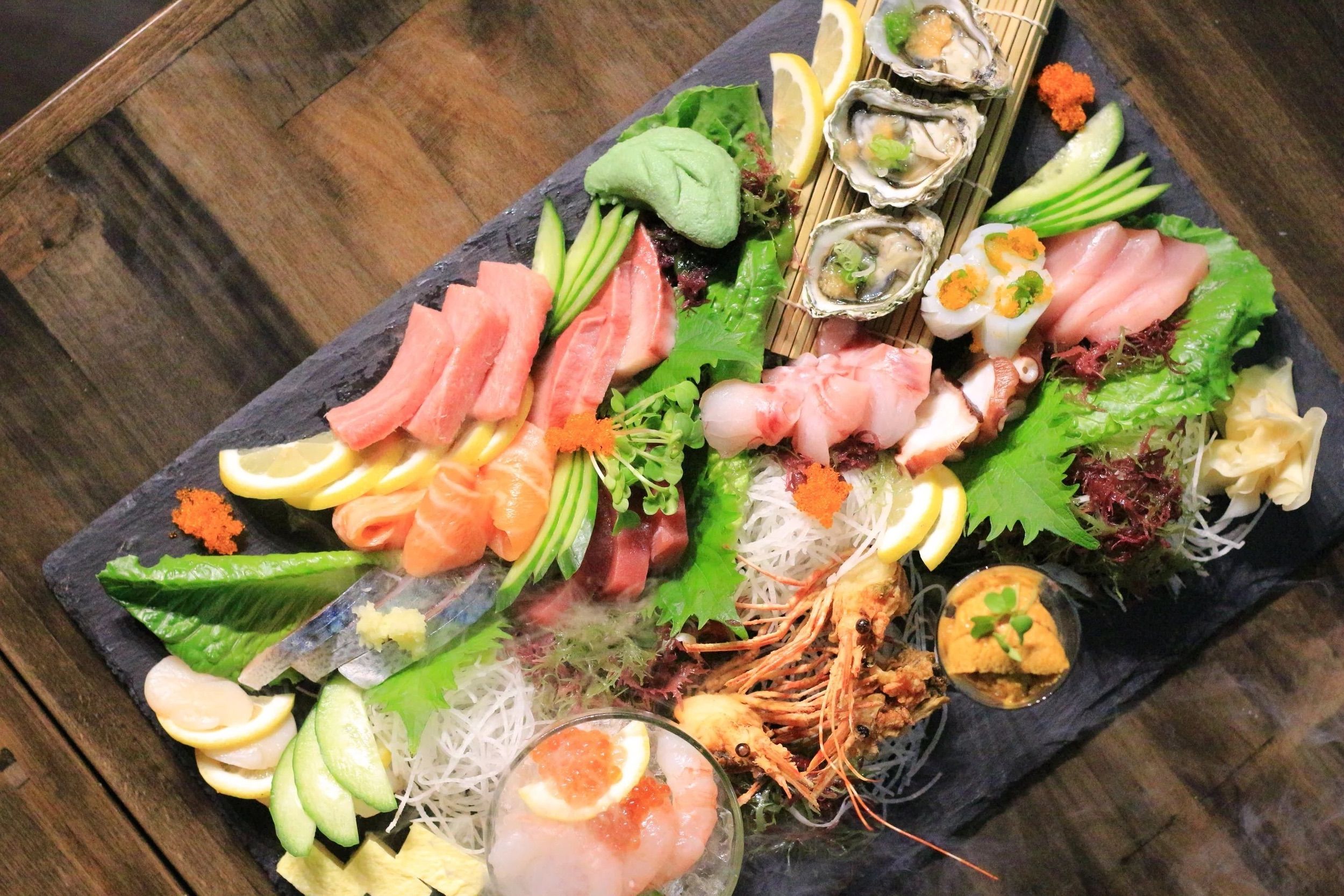 Vibrant Japanese sashimi platter on a slate board with assorted seafood — tuna and salmon slices, oysters, octopus, shrimp heads, uni and ikura roe, avocado slices, lemon wedges and shredded daikon garnish