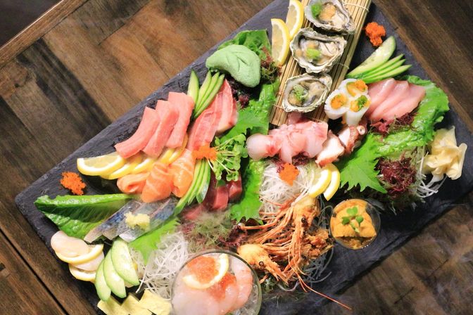 Vibrant Japanese sashimi platter on a slate board with assorted seafood — tuna and salmon slices, oysters, octopus, shrimp heads, uni and ikura roe, avocado slices, lemon wedges and shredded daikon garnish