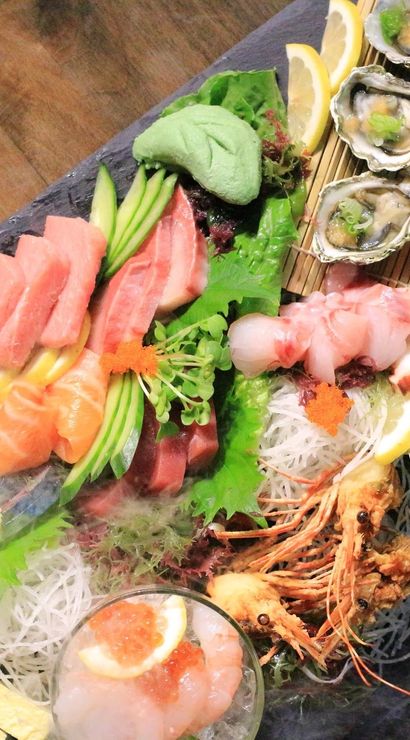 Vibrant Japanese sashimi platter on a slate board with assorted seafood — tuna and salmon slices, oysters, octopus, shrimp heads, uni and ikura roe, avocado slices, lemon wedges and shredded daikon garnish