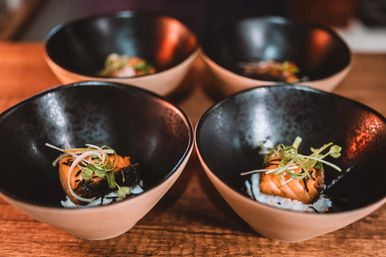 Four ceramic bowls on a wooden table, each holding a seared salmon bite on rice topped with microgreens — Japanese-style sushi appetizer in black-and-beige bowls.