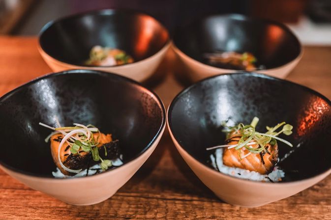 Four ceramic bowls on a wooden table, each holding a seared salmon bite on rice topped with microgreens — Japanese-style sushi appetizer in black-and-beige bowls.