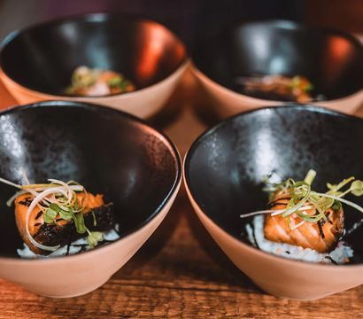 Four ceramic bowls on a wooden table, each holding a seared salmon bite on rice topped with microgreens — Japanese-style sushi appetizer in black-and-beige bowls.