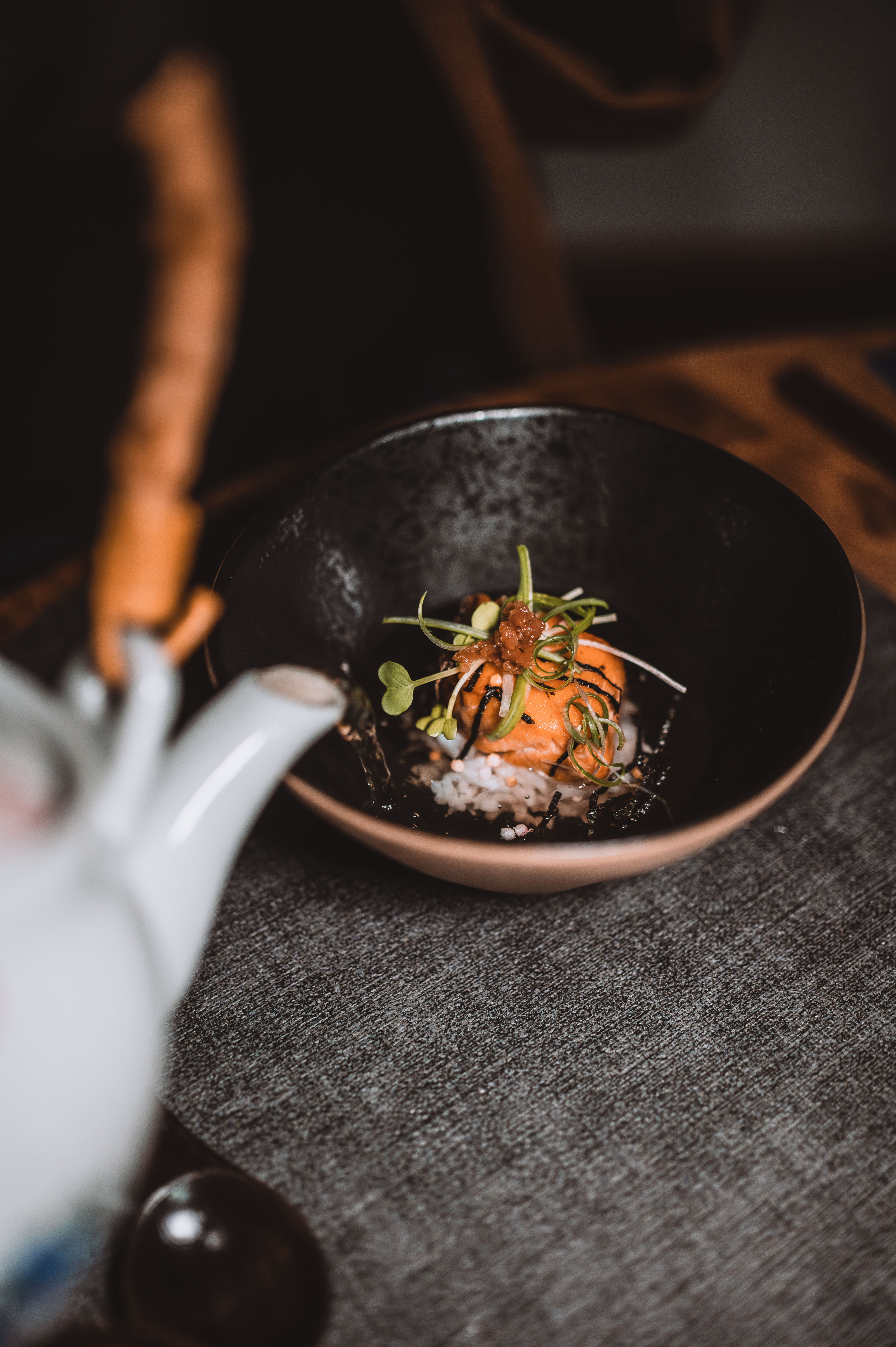 Japanese-style rice bowl with seared salmon and seaweed topped with microgreens, as a white teapot pours warm broth over the rice in a cozy minimalist table setting