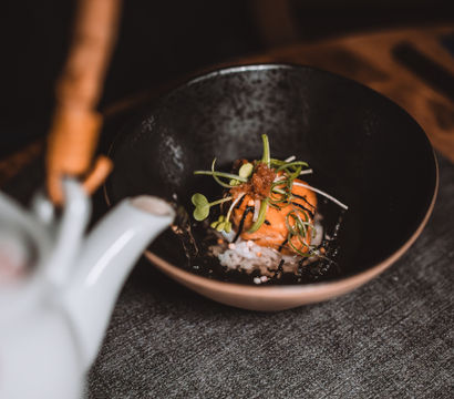 Japanese-style rice bowl with seared salmon and seaweed topped with microgreens, as a white teapot pours warm broth over the rice in a cozy minimalist table setting