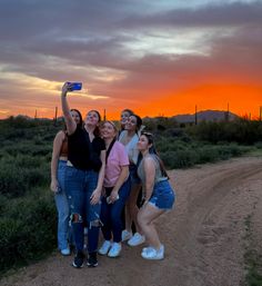 Smiling friends taking a selfie on a desert dirt trail at a vivid orange sunset with saguaro cacti and distant mountains