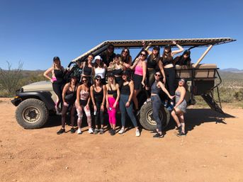 Group of women in athletic outfits posing on and around an open-top off-road vehicle during a sunny desert tour, with cacti and distant mountains under a clear blue sky.