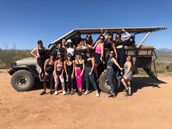 Group of women in athletic outfits posing on and around an open-top off-road vehicle during a sunny desert tour, with cacti and distant mountains under a clear blue sky.
