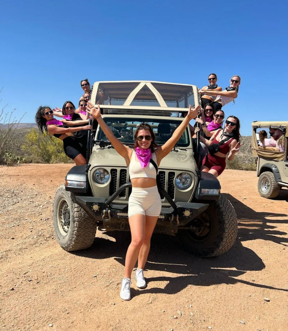 Cheerful group of women in activewear and pink bandanas posing on and around a tan off-road Jeep on a sunny desert trail beneath a clear blue sky