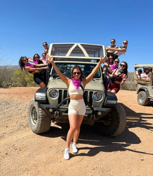 Cheerful group of women in activewear and pink bandanas posing on and around a tan off-road Jeep on a sunny desert trail beneath a clear blue sky