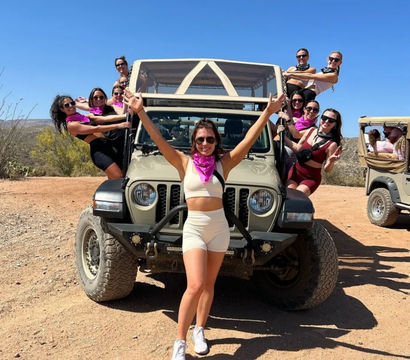 Cheerful group of women in activewear and pink bandanas posing on and around a tan off-road Jeep on a sunny desert trail beneath a clear blue sky