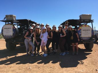 Group of women in bucket hats posing between two off-road jeeps with 'Bachelorette Party' signs on a sunny desert jeep tour, holding drinks and smiling under a clear blue sky.