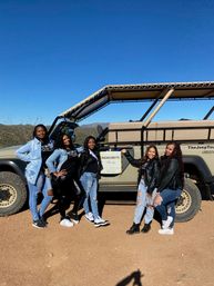 Five friends smiling and posing by an open-air tour jeep with a 'Bachelorette Party' sign in a sunny Southwestern desert landscape with clear blue sky and distant mountains.