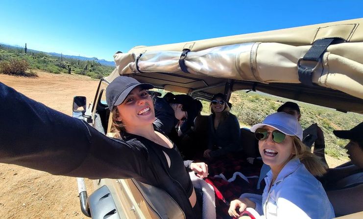 Smiling friends taking a sunny selfie from an open-top jeep driving through a saguaro-studded Sonoran Desert landscape
