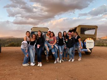 Group of women posing in front of two canvas-top jeeps at a dusty mountain overlook during sunset, casual outfits and ripped jeans, bachelorette party sign on a jeep spare tire, rolling hills and dramatic clouds in the background