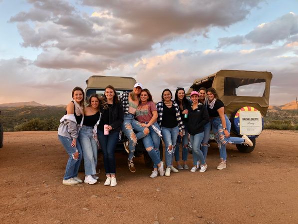 Group of women posing in front of two canvas-top jeeps at a dusty mountain overlook during sunset, casual outfits and ripped jeans, bachelorette party sign on a jeep spare tire, rolling hills and dramatic clouds in the background