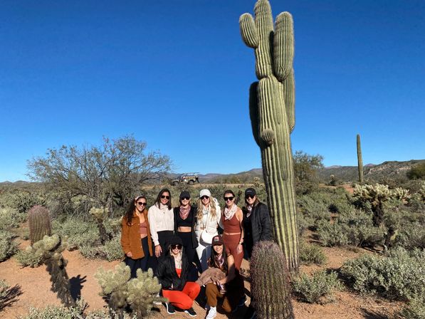 Eight people posing beside a towering saguaro cactus in the sunny Sonoran Desert in Arizona, surrounded by low desert shrubs, distant hills and a clear blue sky.