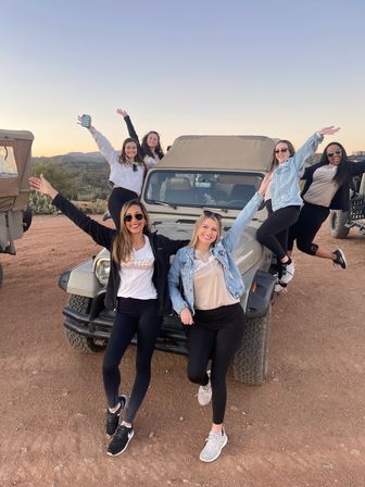 Six women posing and cheering on and around a tan off-road jeep on a red-dirt desert trail at sunset, arms raised and smiling with cacti and distant mountains in the background.