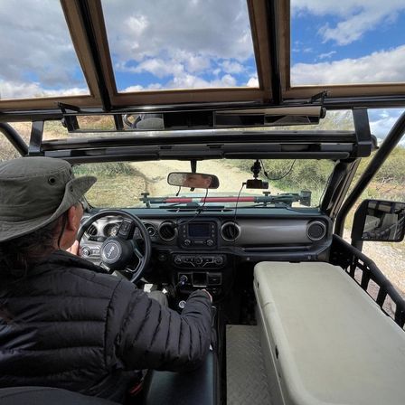 Open-top Jeep on a desert off-road trail, view from the back seat showing dashboard, cooler, and a hat-wearing driver steering under a blue sky with clouds.