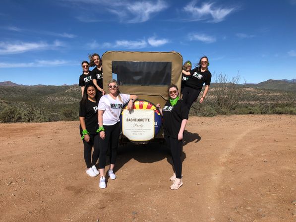 Group of women at a bachelorette party posing around a rugged off-road vehicle with a bachelorette sign on the spare tire, standing on a sunlit desert dirt road with scrubby hills and a bright blue sky.