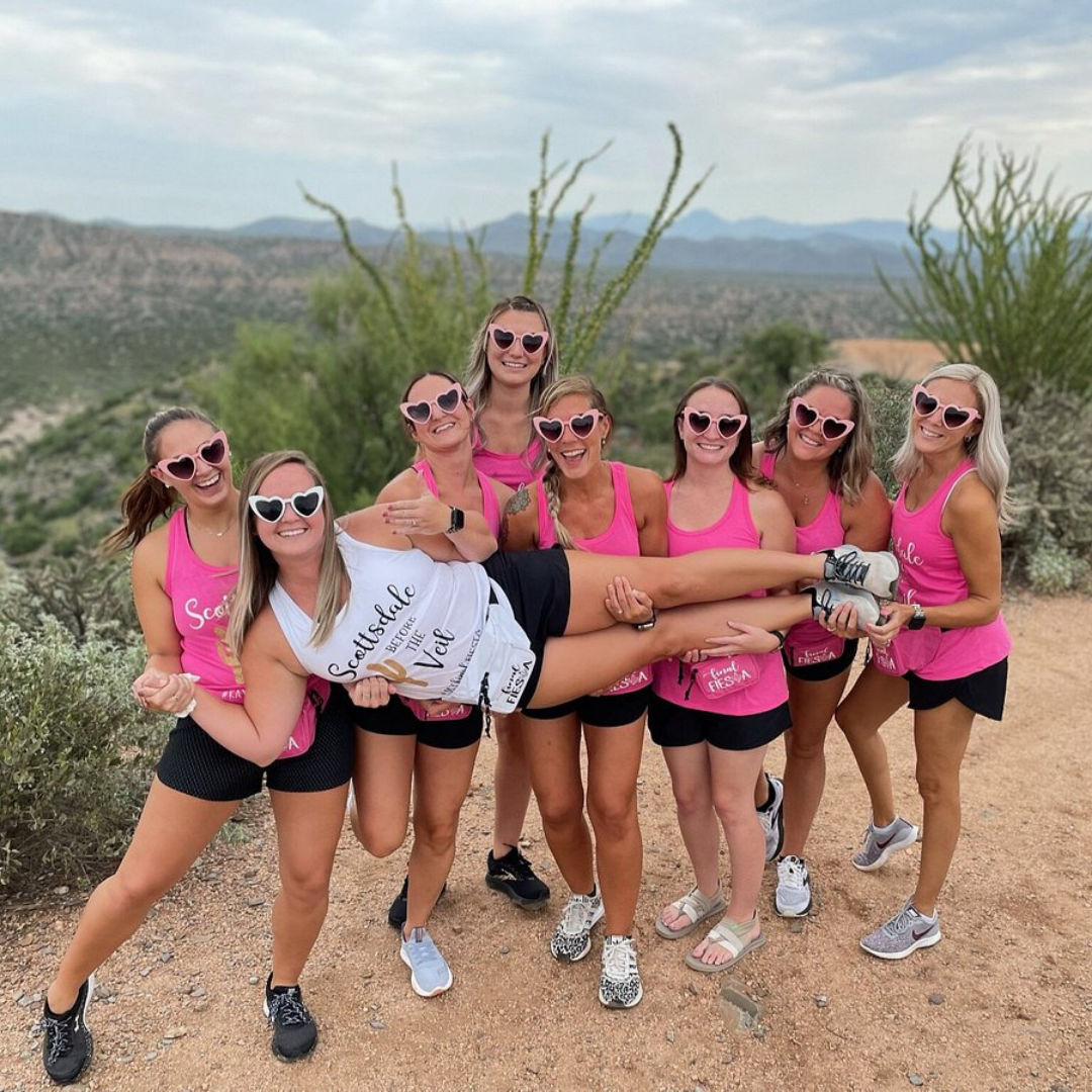 Group of eight women in pink tank tops and heart-shaped sunglasses on a desert hiking trail with cacti and distant mountains, laughing as they hold a friend horizontally in a playful celebration.