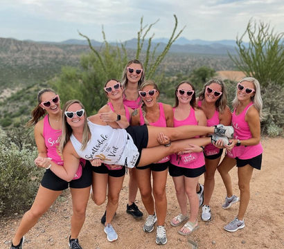 Group of eight women in pink tank tops and heart-shaped sunglasses on a desert hiking trail with cacti and distant mountains, laughing as they hold a friend horizontally in a playful celebration.