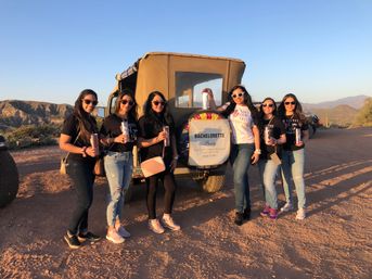 Seven women in sunglasses posing with tumblers beside a rugged off-road vehicle displaying a 'Bachelorette Party' sign, set on a sunlit Southwestern desert road with cacti and distant mountains at sunset.