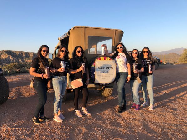 Seven women in sunglasses posing with tumblers beside a rugged off-road vehicle displaying a 'Bachelorette Party' sign, set on a sunlit Southwestern desert road with cacti and distant mountains at sunset.