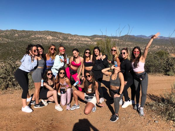 Energetic group of women in colorful activewear posing on a sunny desert hiking trail with rugged mountain ridges and a clear blue sky, some holding water bottles and smiling.