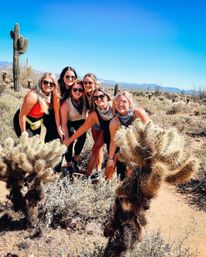 Six friends in sunglasses and bandanas smiling and posing among saguaro and cholla cacti in a sunny Sonoran desert under a clear blue sky with distant mountains.