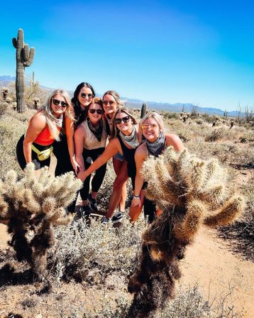 Six friends in sunglasses and bandanas smiling and posing among saguaro and cholla cacti in a sunny Sonoran desert under a clear blue sky with distant mountains.