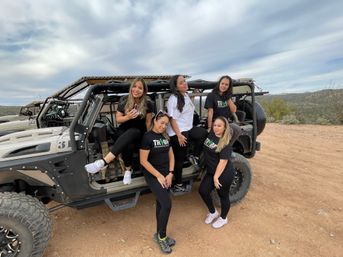 Five smiling women in matching shirts posing on and around a rugged off-road vehicle on a desert trail with cloudy skies and distant hills — outdoor off-road adventure.