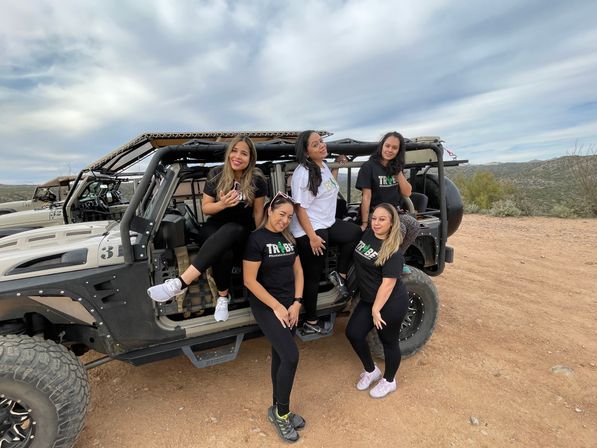 Five smiling women in matching shirts posing on and around a rugged off-road vehicle on a desert trail with cloudy skies and distant hills — outdoor off-road adventure.