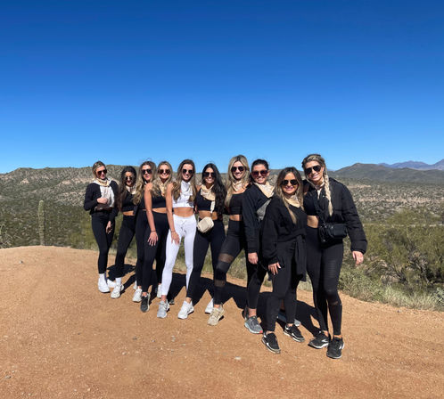 Smiling group of women in activewear posing on a sunlit desert hiking trail with cacti and rugged mountain vistas under a clear blue sky — outdoor adventure vibe.