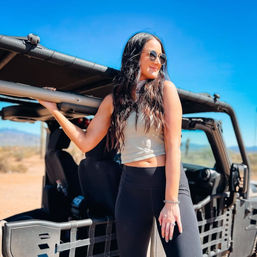 Smiling woman in sunglasses leaning on an open-top 4x4 in a sunlit desert under a clear blue sky, wearing a cropped tank and black leggings.