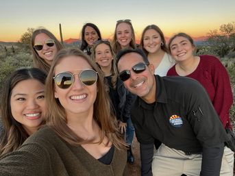 Smiling group selfie of nine adults wearing casual clothes and sunglasses at golden-hour in the Arizona desert, with a saguaro cactus and distant mountains at sunset in the background.