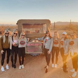Group of seven friends in matching bachelorette shirts posing by a tour jeep at sunset in the Sonoran Desert with saguaro cacti in the background — fun Arizona desert celebration.