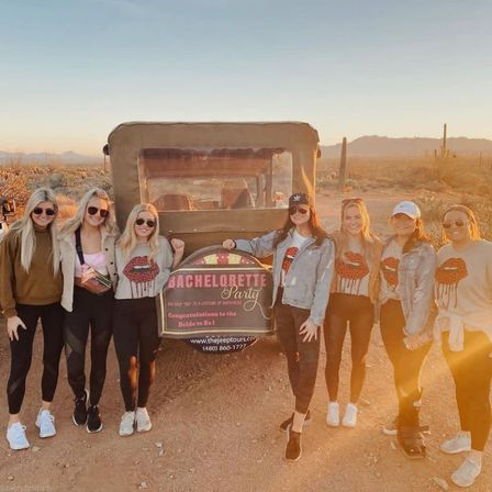 Group of seven friends in matching bachelorette shirts posing by a tour jeep at sunset in the Sonoran Desert with saguaro cacti in the background — fun Arizona desert celebration.
