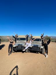 Group of friends posing on and around two tan jeeps on a sunny desert trail, arms raised under a vivid blue sky with cactus-studded hills in the background