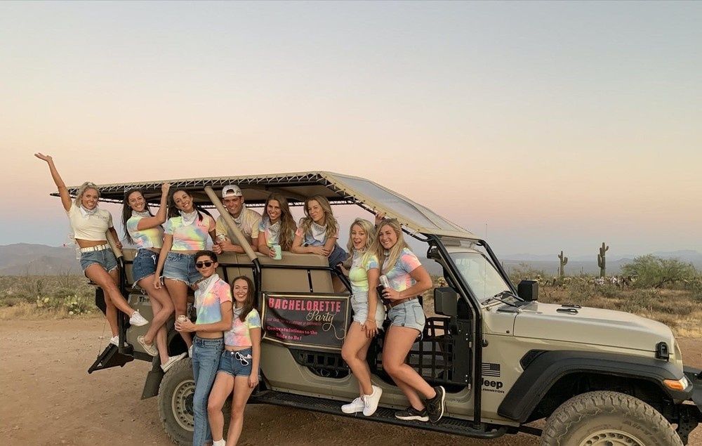 Group of friends in tie-dye shirts posing on an open-air tour Jeep at sunset in a desert with saguaro cacti for a bachelorette party.