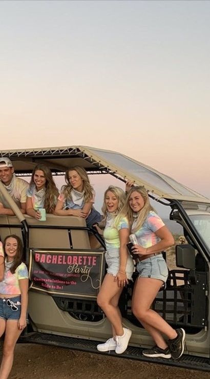Group of friends in tie-dye shirts posing on an open-air tour Jeep at sunset in a desert with saguaro cacti for a bachelorette party.
